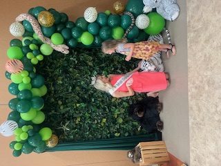 A child talks with a woman in a pink dress in front of a leafy backdrop and green balloons.
