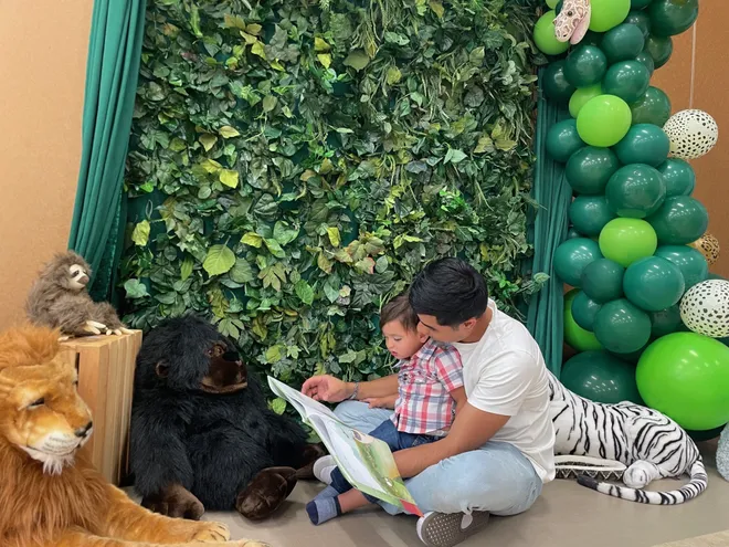 An adult and child read a picture book together beside plush animals and a leafy backdrop.